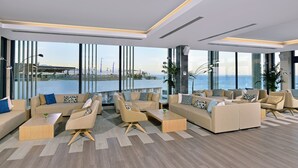 Un área de descanso moderna con varios sofás y sillones, ventanas amplias con vistas al océano y piso de madera.