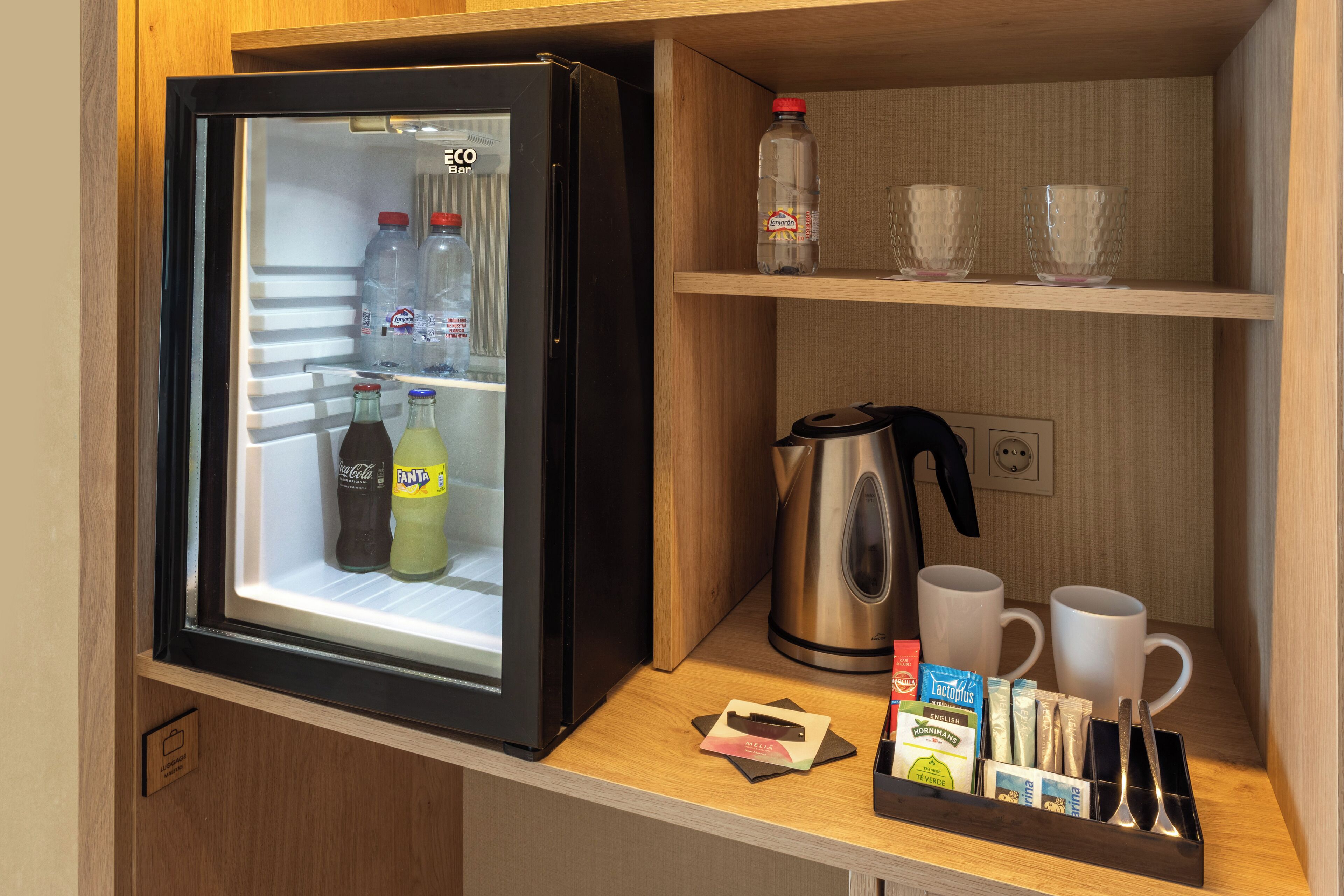 Ein kompakter Kühlschrank mit Getränken, einem Wasserkocher und einer Kaffeemaschine auf einer Holzauslage.