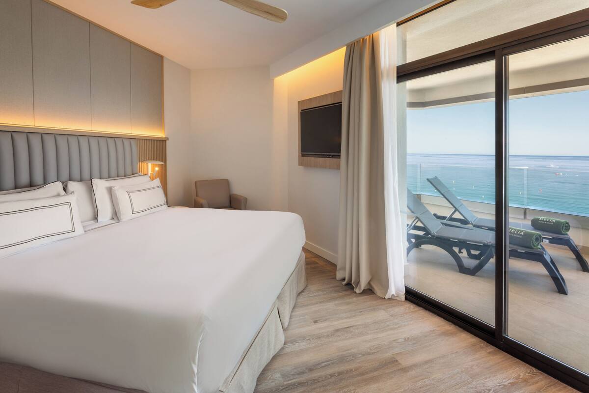 Ett hotellrum med en stor säng, en tv som är monterad på väggen och en balkong med utsikt över havet.