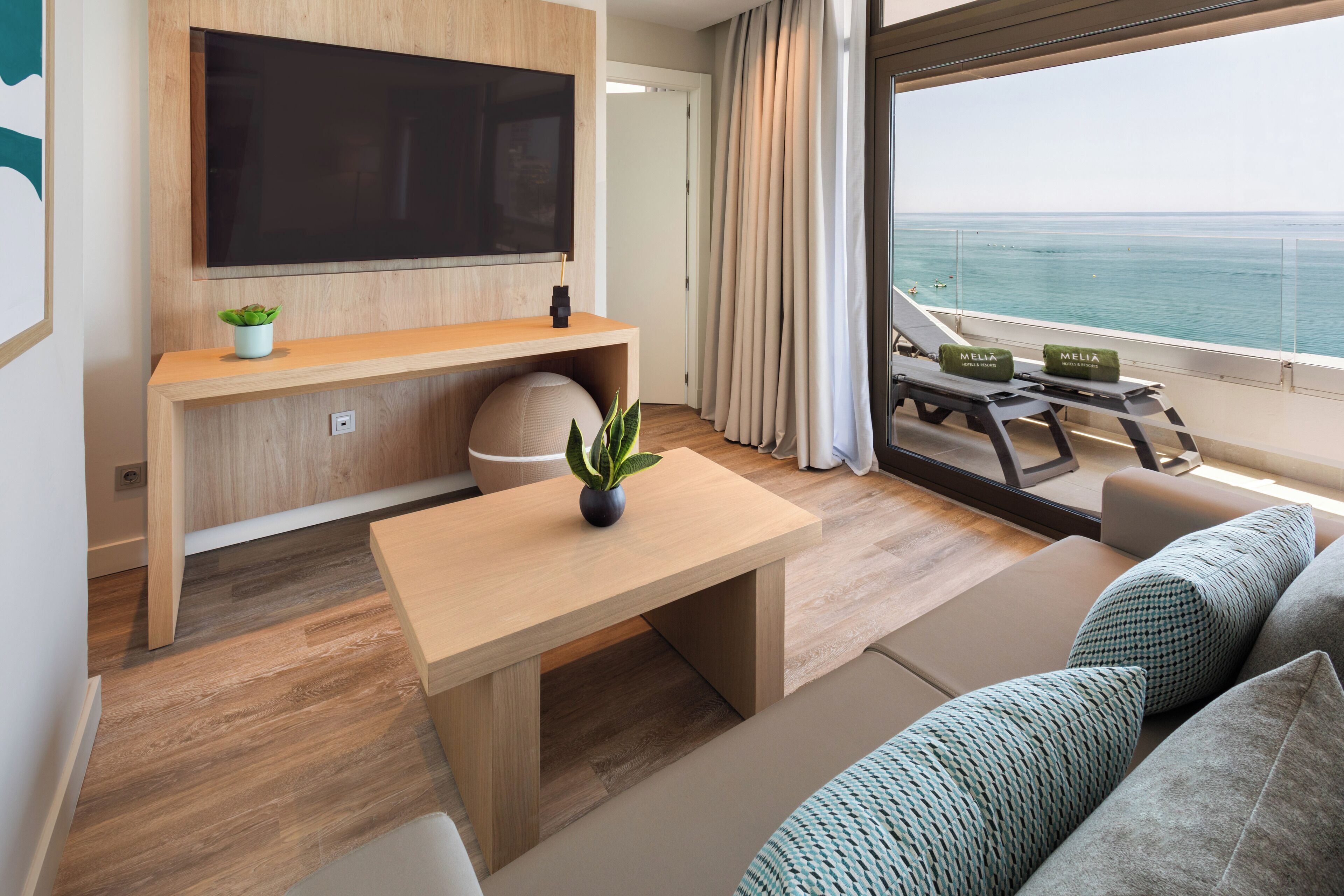 Una sala de estar moderna con televisor de pantalla plana, muebles de madera, una planta pequeña y un balcón con vista al mar.