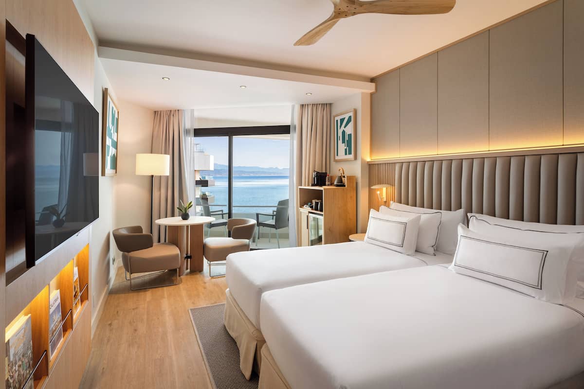Ett hotellrum med två sängar, en TV, ett skrivbord och utsikt över havet.