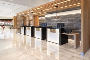 Zona de recepción con tres mostradores, cada uno con una computadora y un letrero que dice “MEDIA”. El espacio presenta un diseño moderno, con listones de madera en el techo y una pared de piedra detrás de los mostradores.