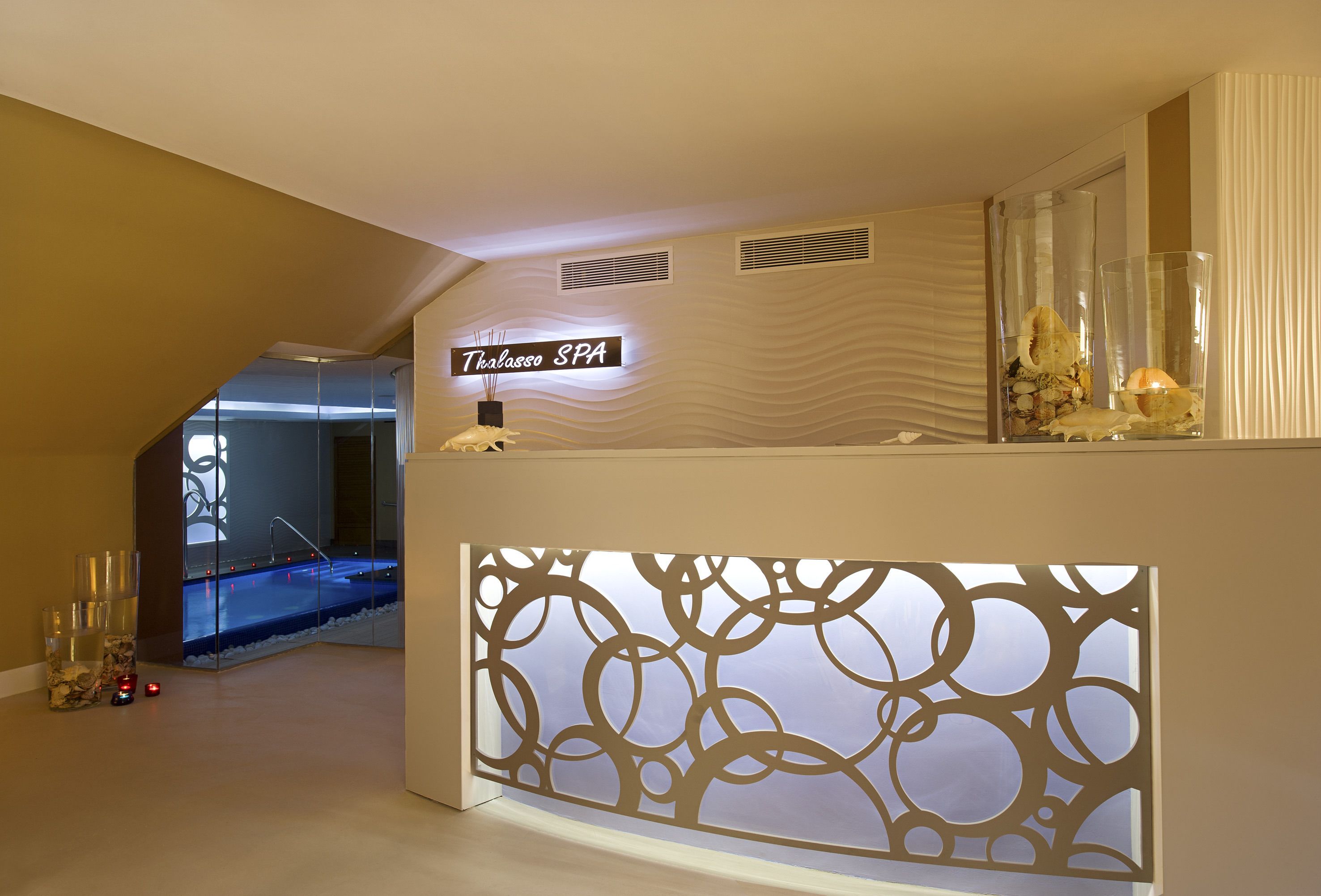 Ein Empfangsbereich mit einem Schild, auf dem "Thalasso SPA" steht, einer Wand mit dekorativen Kreismustern und einer Glasvitrine mit Muscheln.