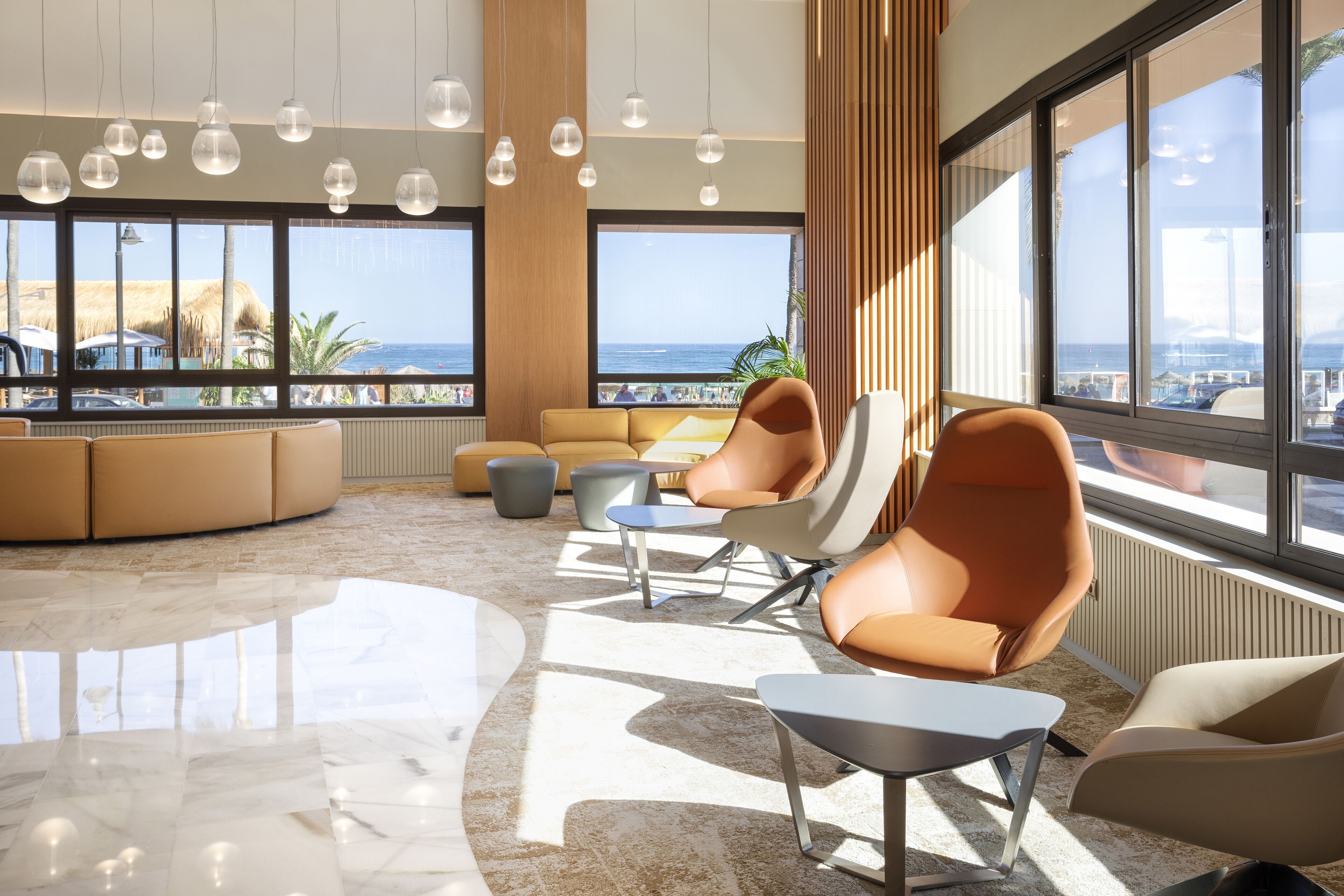 Un salón moderno con un sofá curvo, una mesa de centro y sillas naranjas. Amplios ventanales ofrecen vista a la playa y al océano.