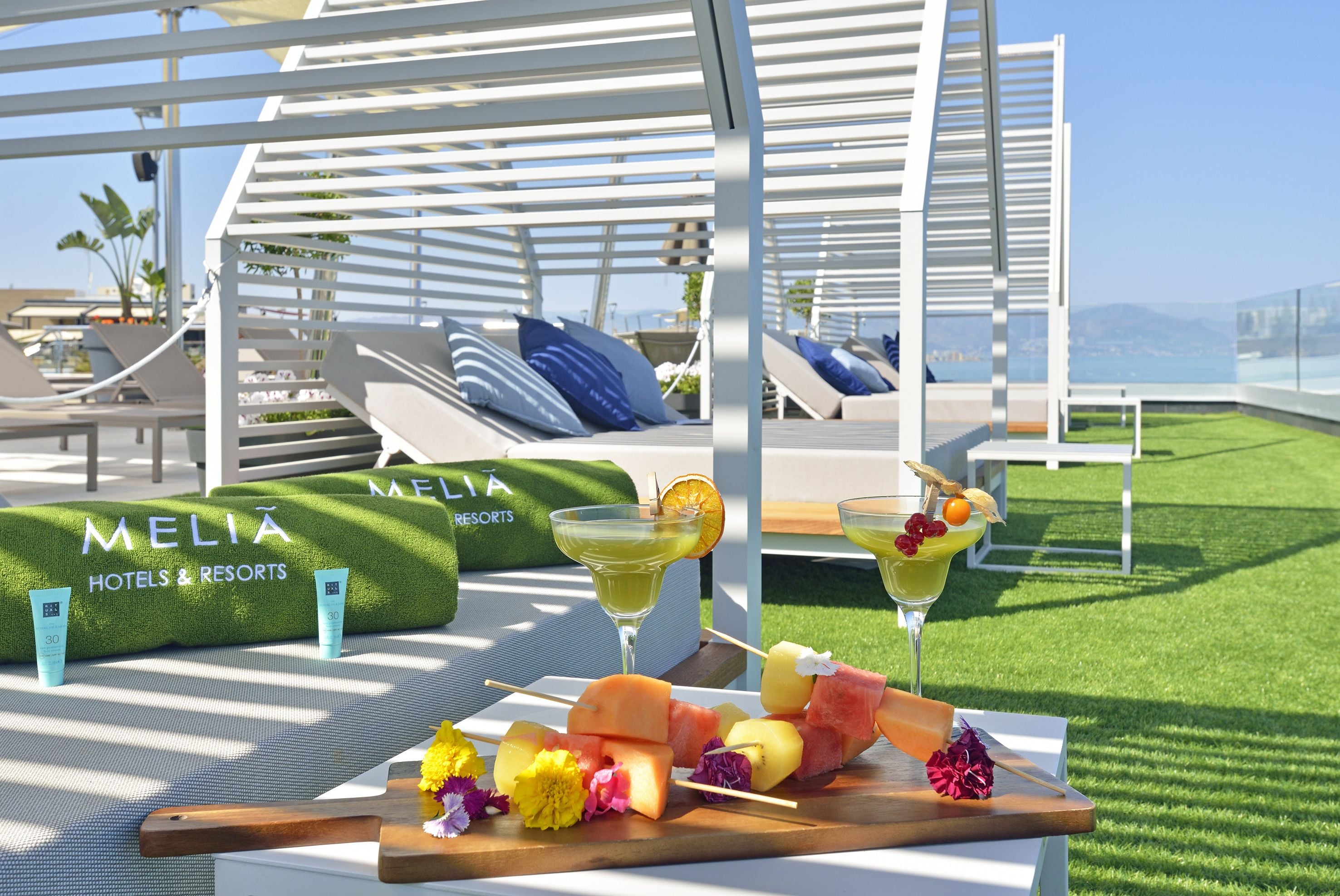 Un área de descanso junto a la piscina con sillones reclinables, una mesa de madera con brochetas de fruta y bebidas, y una zona cubierta de césped artificial con un letrero de 'MELIA HOTELS & RESORTS'.