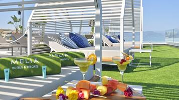 Un área de descanso junto a la piscina con sillones reclinables, una mesa de madera con brochetas de fruta y bebidas, y una zona cubierta de césped artificial con un letrero de 'MELIA HOTELS & RESORTS'.