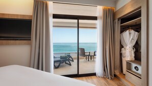 Habitación de hotel con una cama grande, puerta de vidrio corrediza a un balcón con vista al mar, zona de estar con una mesa y sillas, y armario con batas.