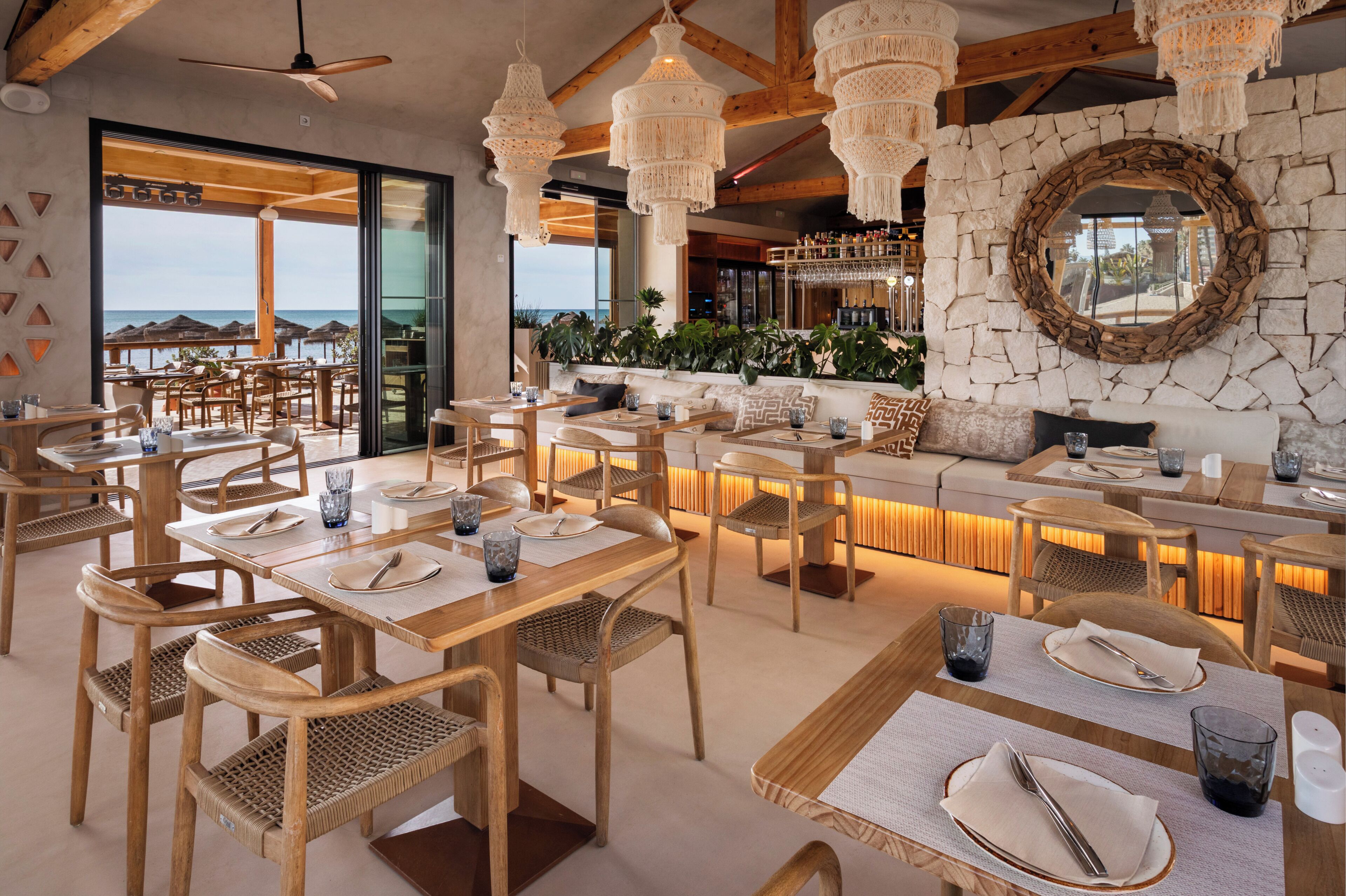 Un restaurante con mesas y sillas de madera, una pared de piedra con un espejo y una zona de bar con plantas.