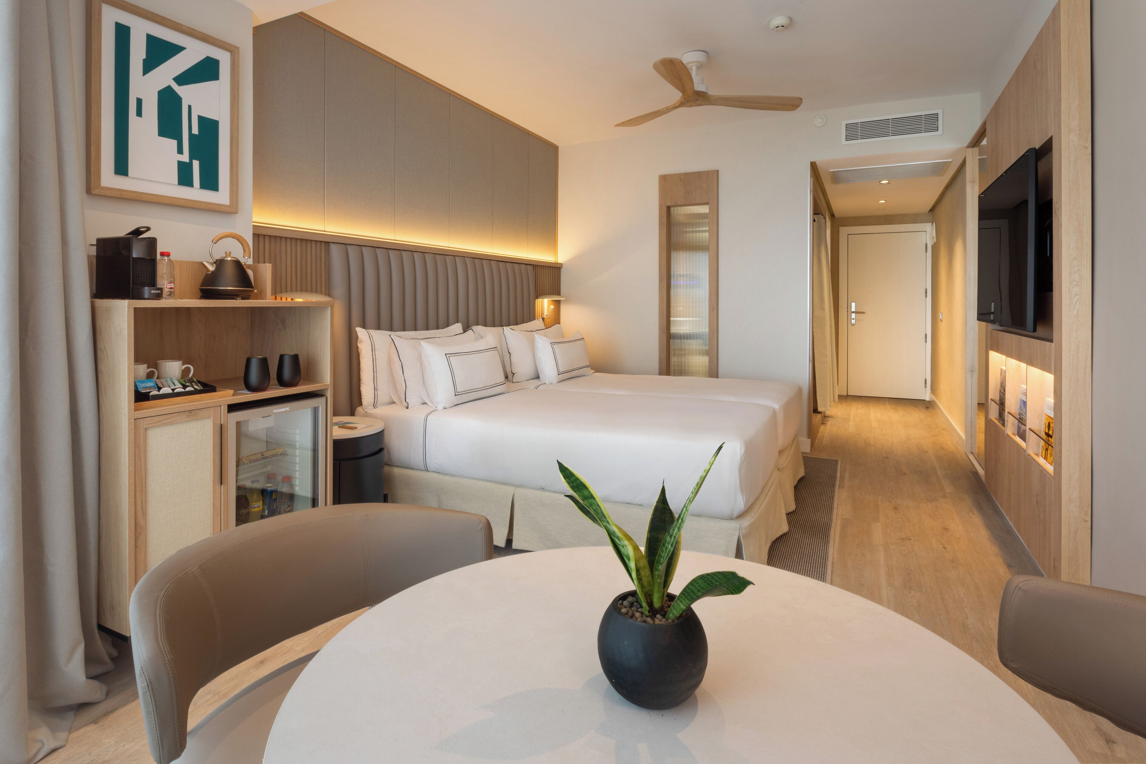 Ein modernes Hotelzimmer mit einem Bett, einem Schreibtisch mit Stuhl, einem kleinen Tisch mit Pflanze, einem Fernseher und einem Deckenventilator.