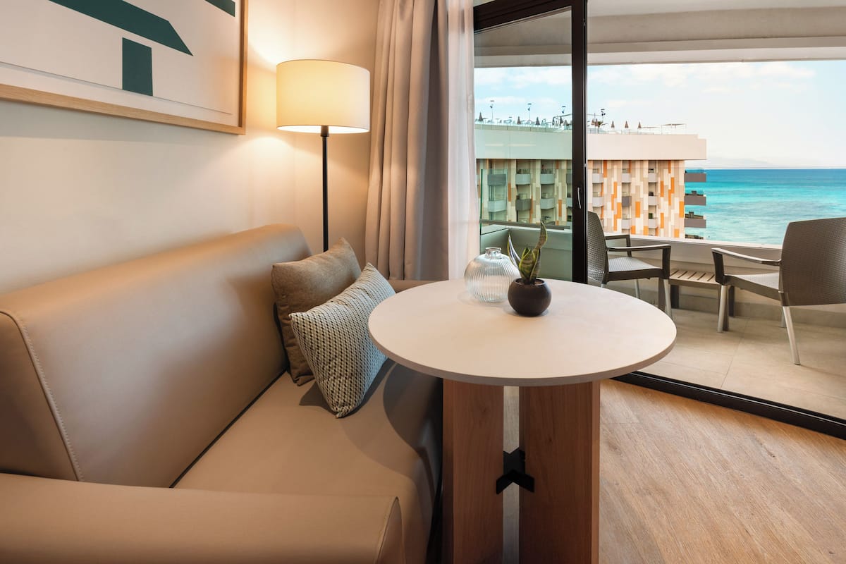 Ett hotellrum med en soffa, ett bord med en vas och utsikt över havet och byggnaderna.