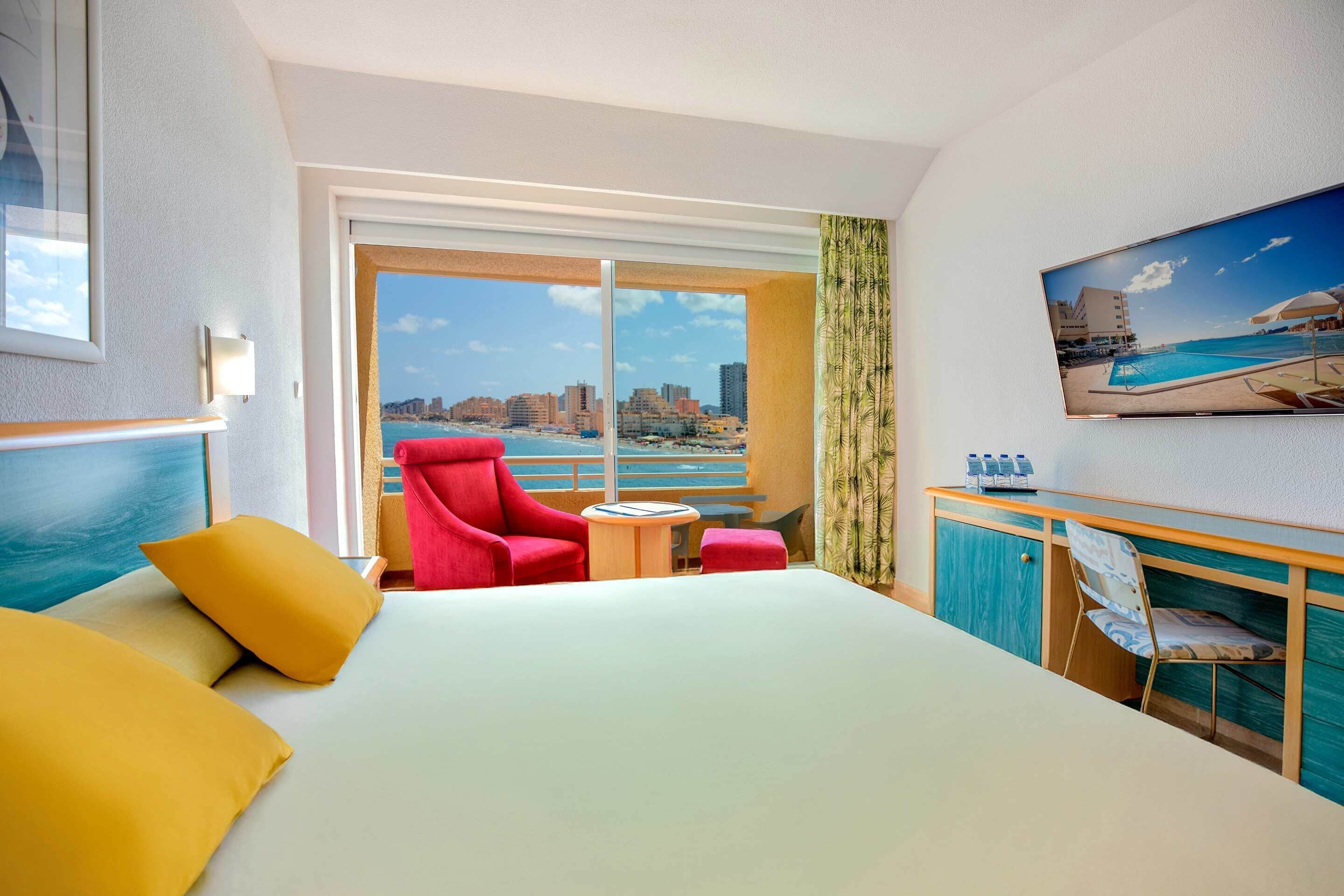 A hotel room with a bed, a red chair, a small table, a TV, and a large window with a city view.