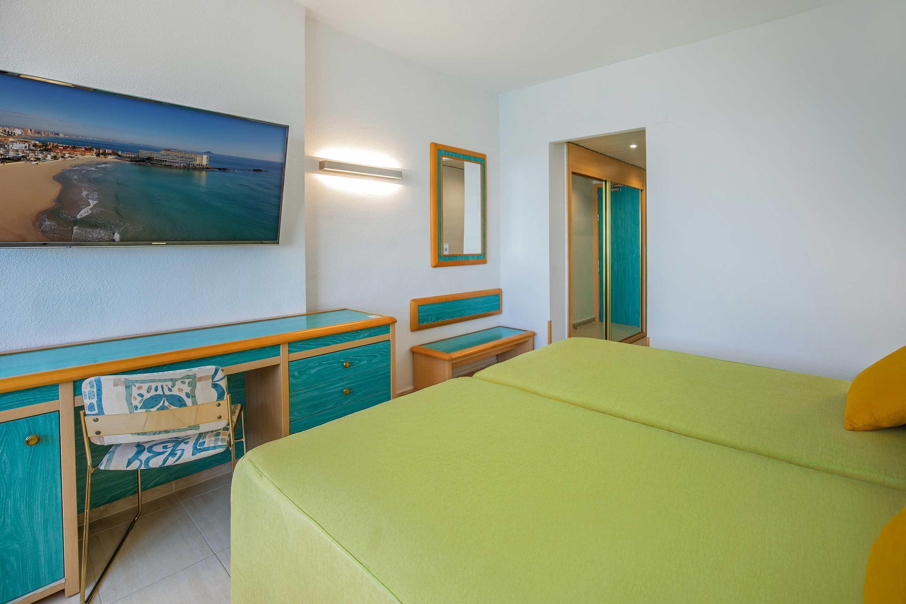 A hotel room with a bed, a desk with a chair, a dresser, a mirror, and a painting of a beach scene.