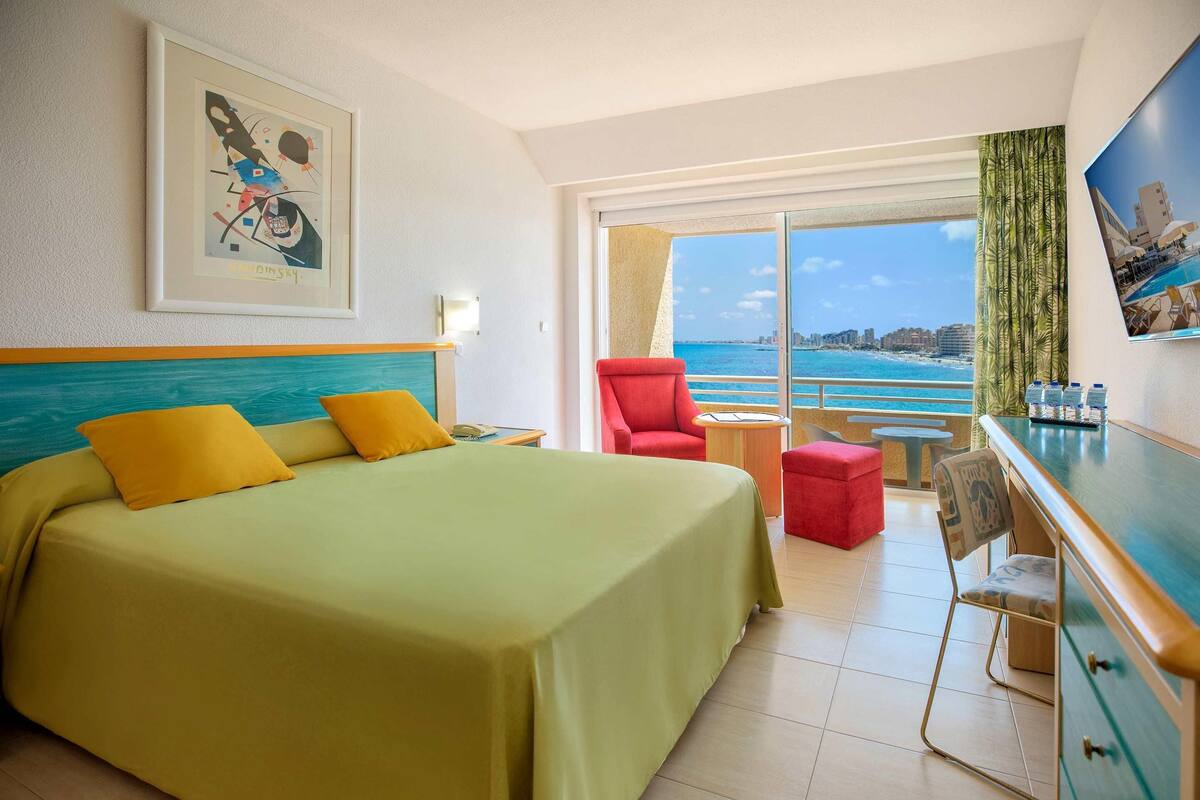 A hotel room with a bed, a red chair, a small table, and a view of the ocean.