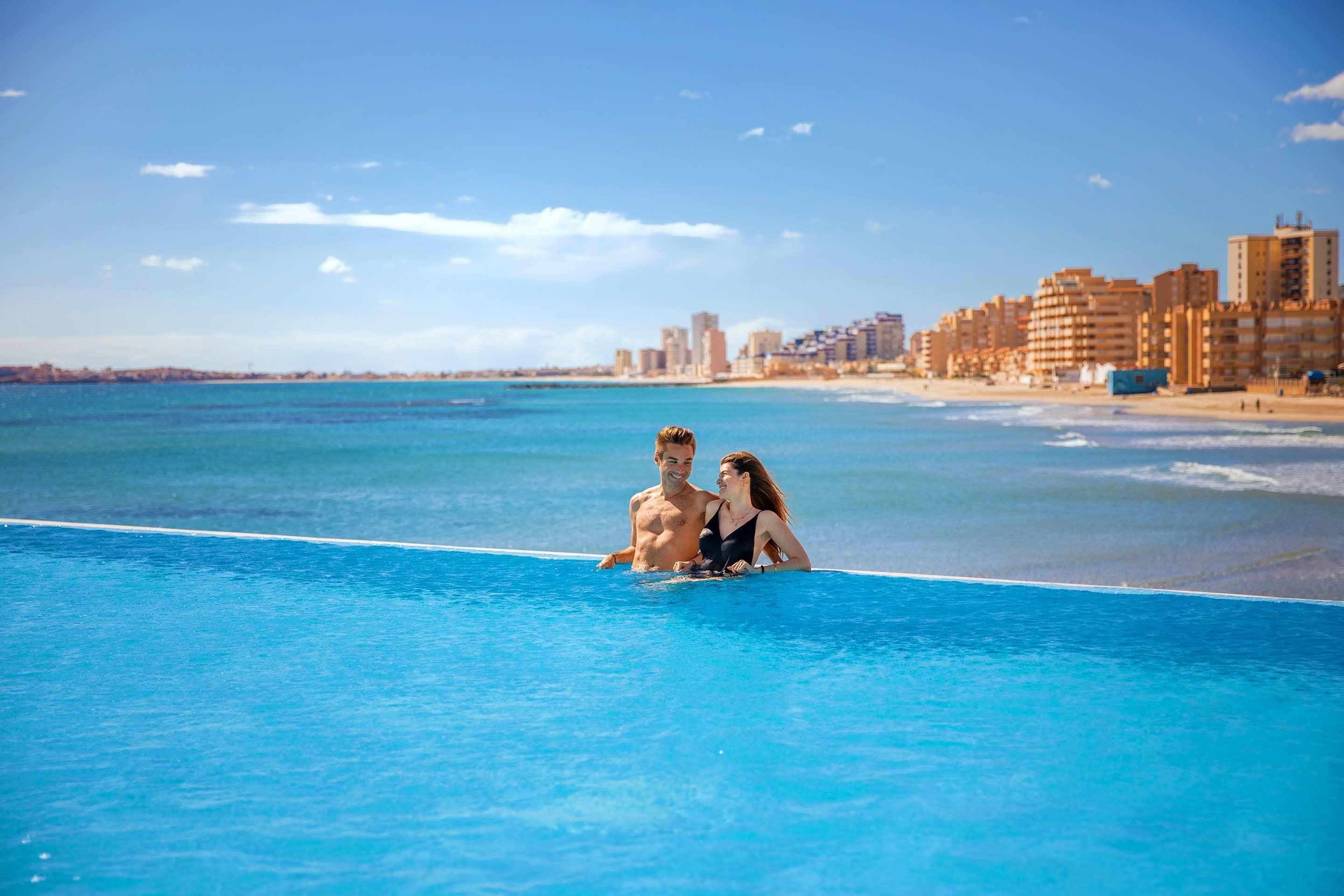 A couple in a swimming pool with a beach and buildings in the background.