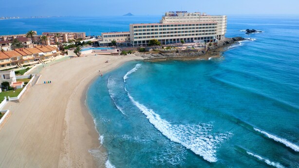 A coastal hotel with multiple floors, a beach, and clear blue waters.