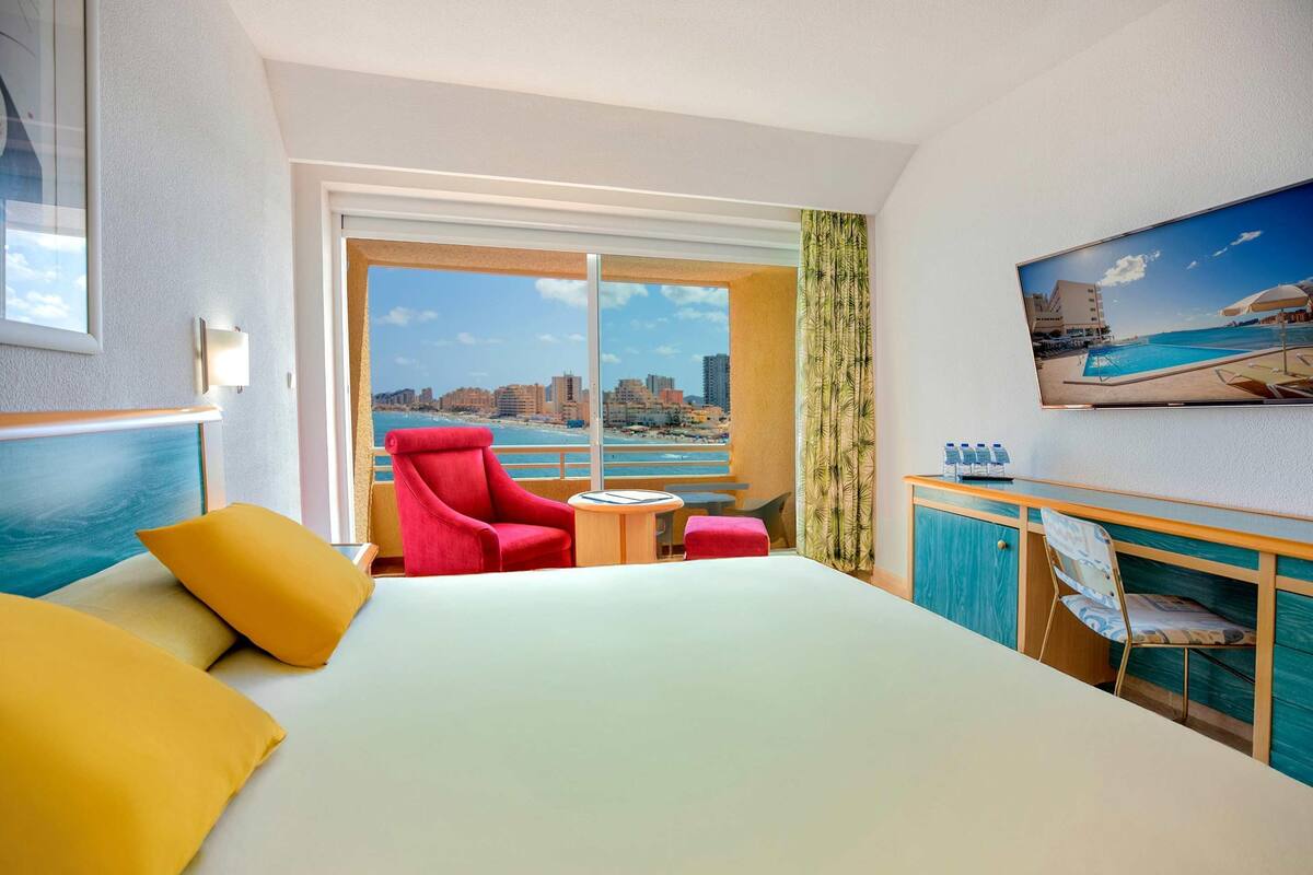 A hotel room with a bed, a red armchair, a small round table, a TV on the wall, and a view of the city and beach.