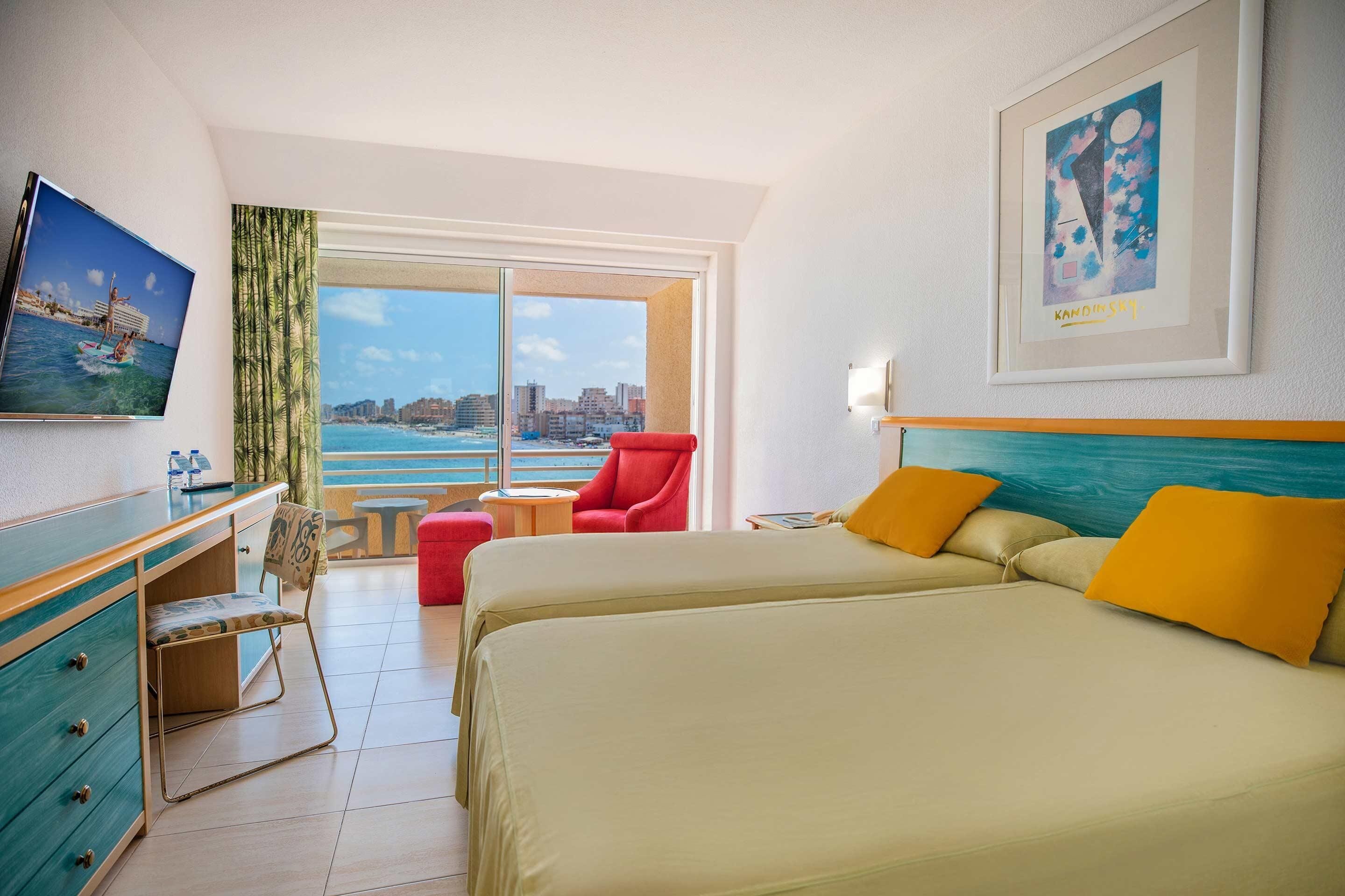 A hotel room with a large bed, a desk with drawers, a chair, a red armchair, a window with a city view, and a framed picture on the wall.