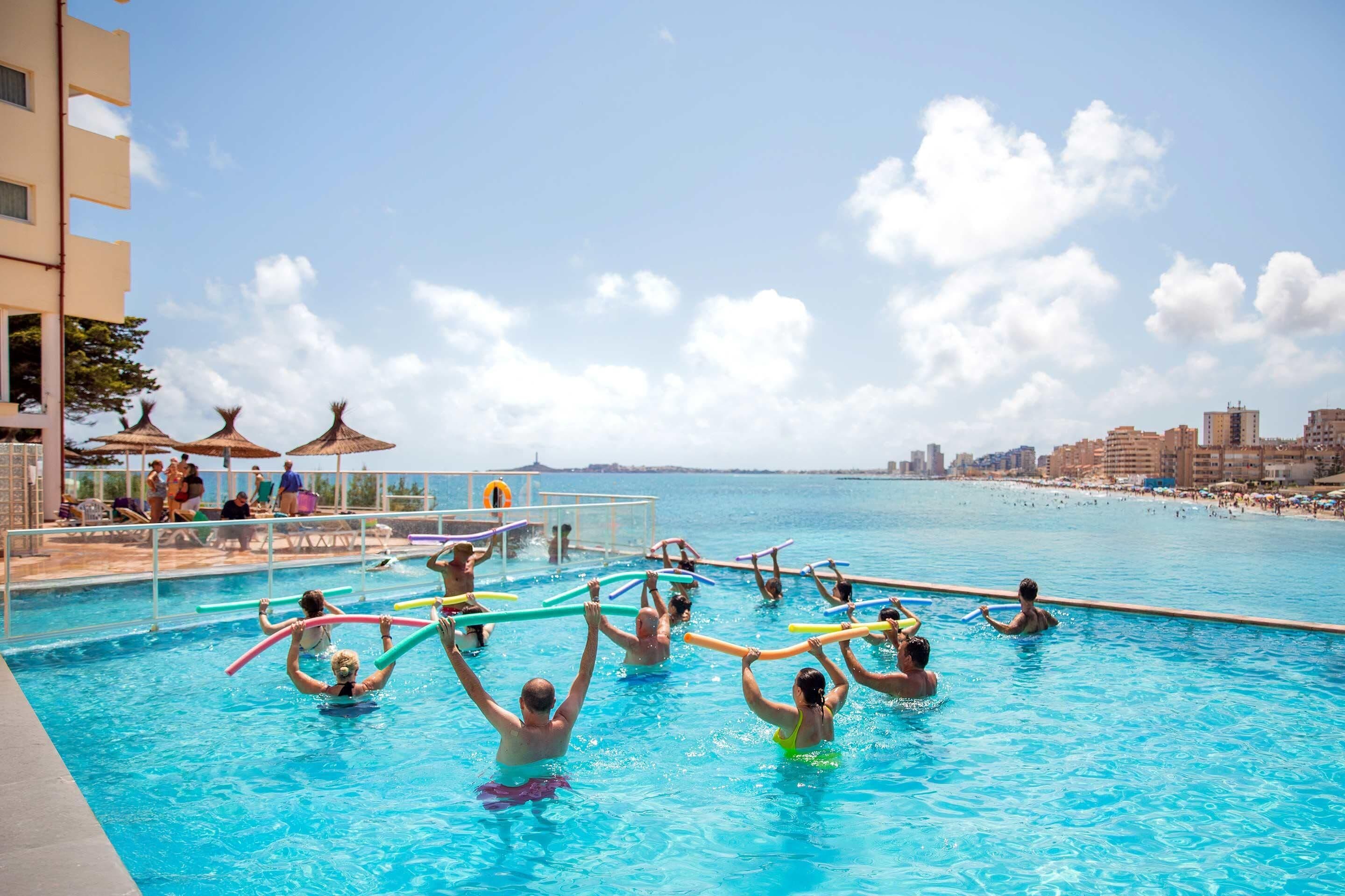 Uma área de piscina com pessoas usando boias, um posto de salva-vidas e vista para a praia e o horizonte da cidade.