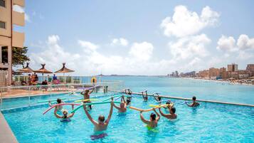 Uma área de piscina com pessoas usando boias, um posto de salva-vidas e vista para a praia e o horizonte da cidade.