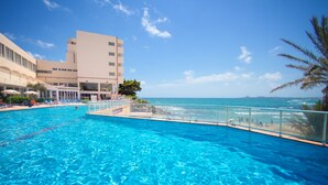 A swimming pool with clear blue water, a beach view, and a building with multiple floors and balconies.