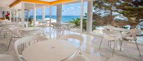 Ein stilvoller Essbereich mit weißen Tischen und Stühlen, große Fenster mit Blick auf das Meer und Bäume.