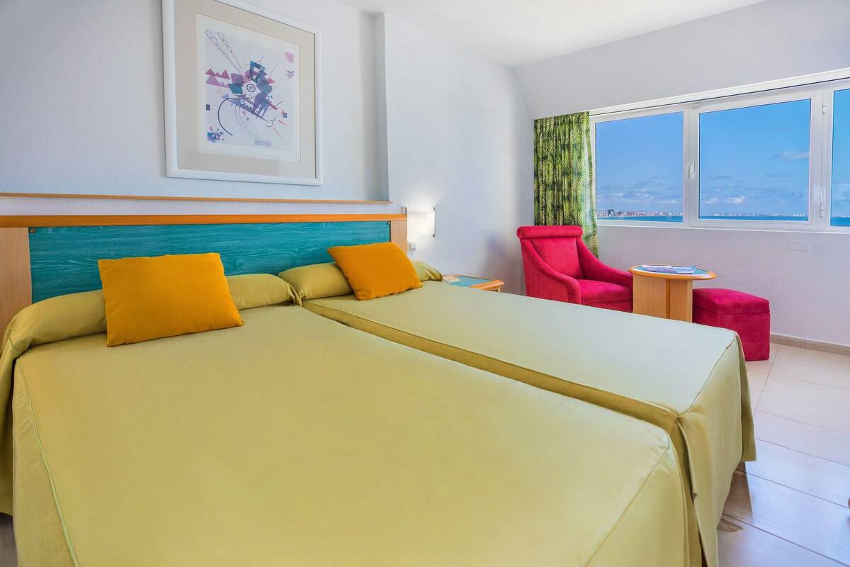 A hotel room with two beds, a red chair, a small table, and a large window with a view.