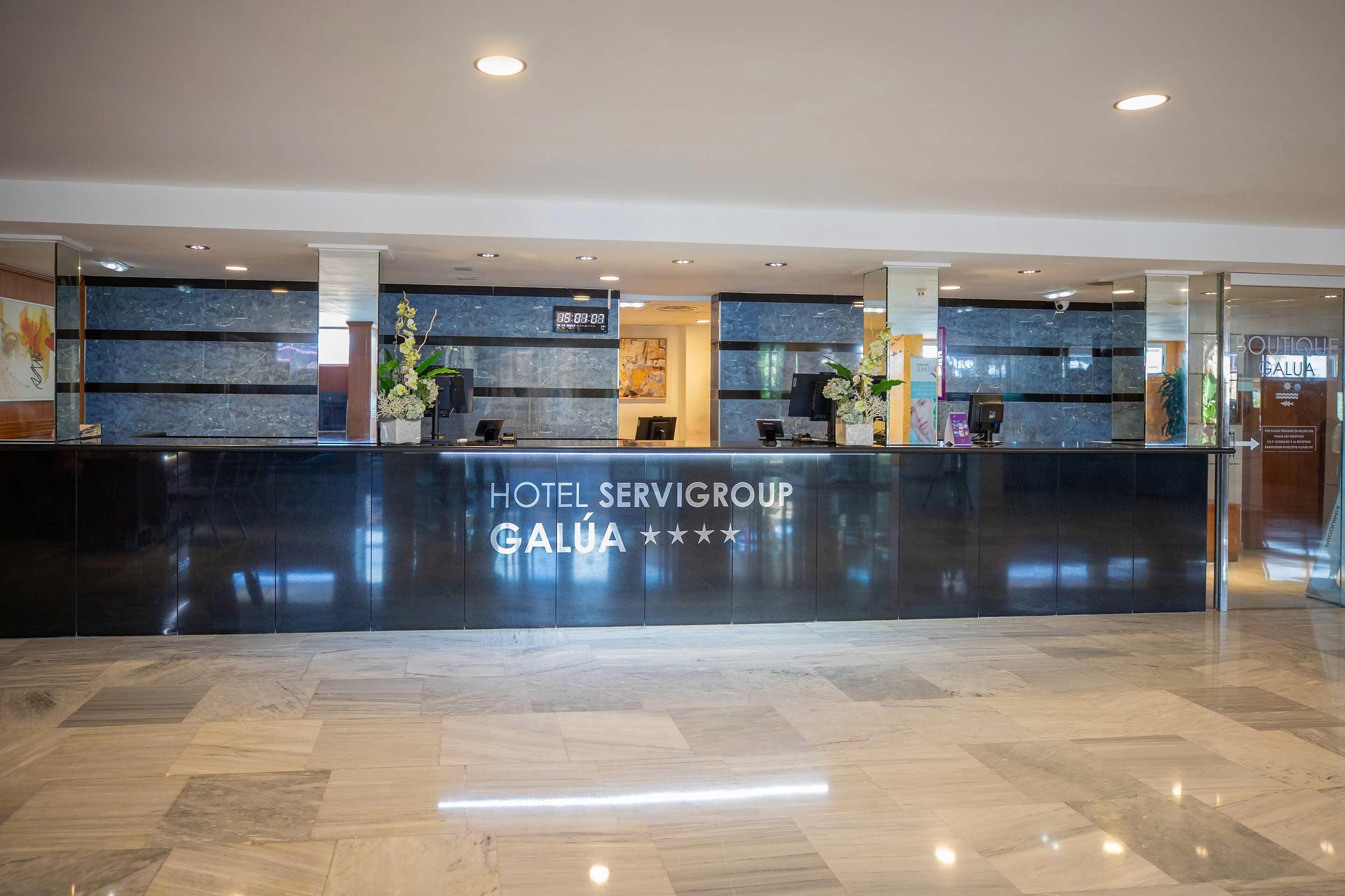 Zona de recepción de hotel con mostrador de mármol, diseño moderno y un reloj digital que muestra la hora.
