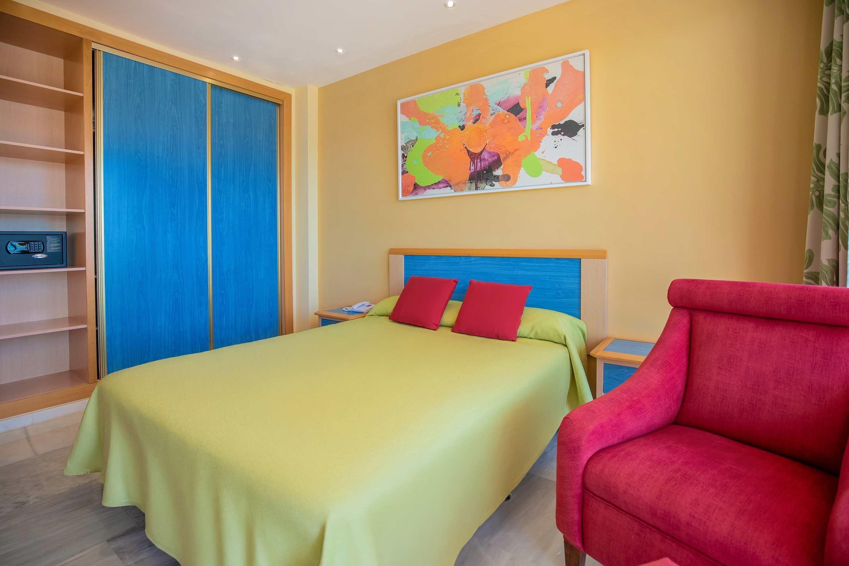 A hotel room with a bed, a red chair, a bookshelf, and a colorful abstract painting on the wall.