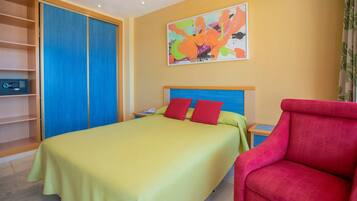 A hotel room with a bed, a red chair, a bookshelf, and a colorful abstract painting on the wall.