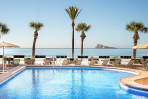 Zona de piscina con sillones de descanso y palmeras, con vista a la playa y el mar.