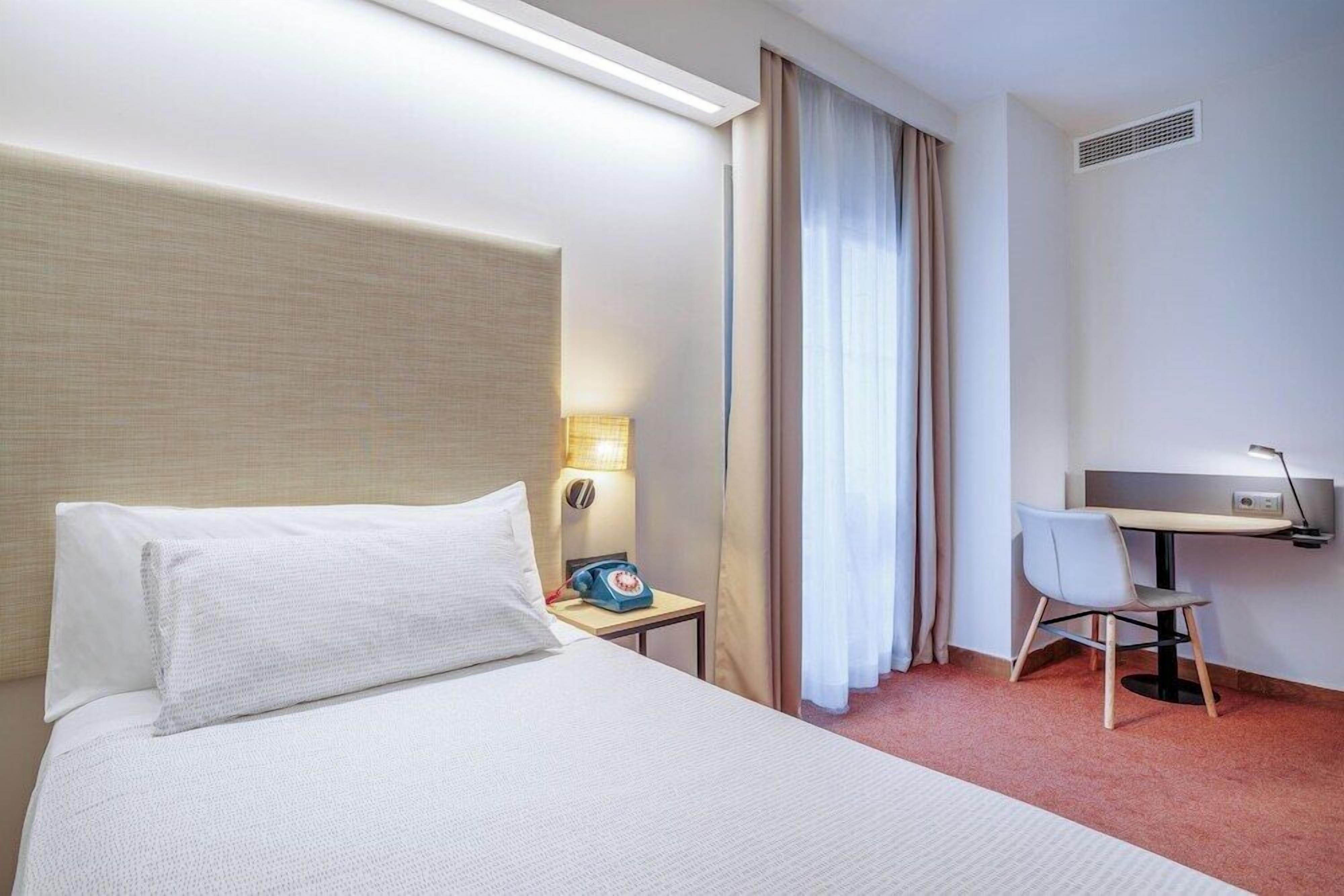 Una habitación de hotel con cama, escritorio, silla, lámpara y teléfono.