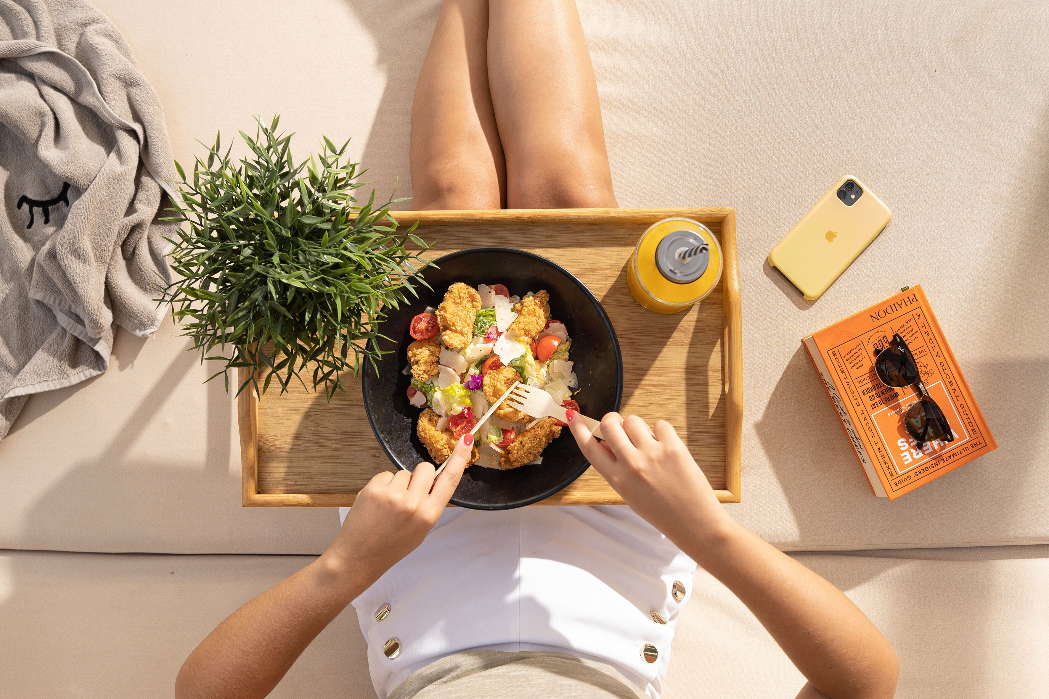 Une personne allongée sur un lit, avec une table d’appoint contenant de la nourriture, une plante, un livre et un téléphone.