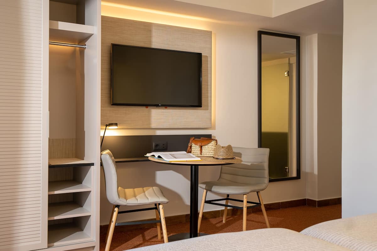 A hotel room with a flat-screen TV mounted on the wall, a small table with a book and a hat, two chairs, and a large mirror.