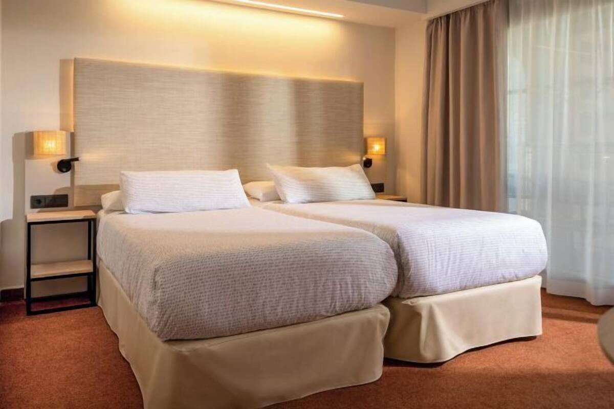 A hotel room with two beds, a nightstand, a lamp, and a window with curtains.
