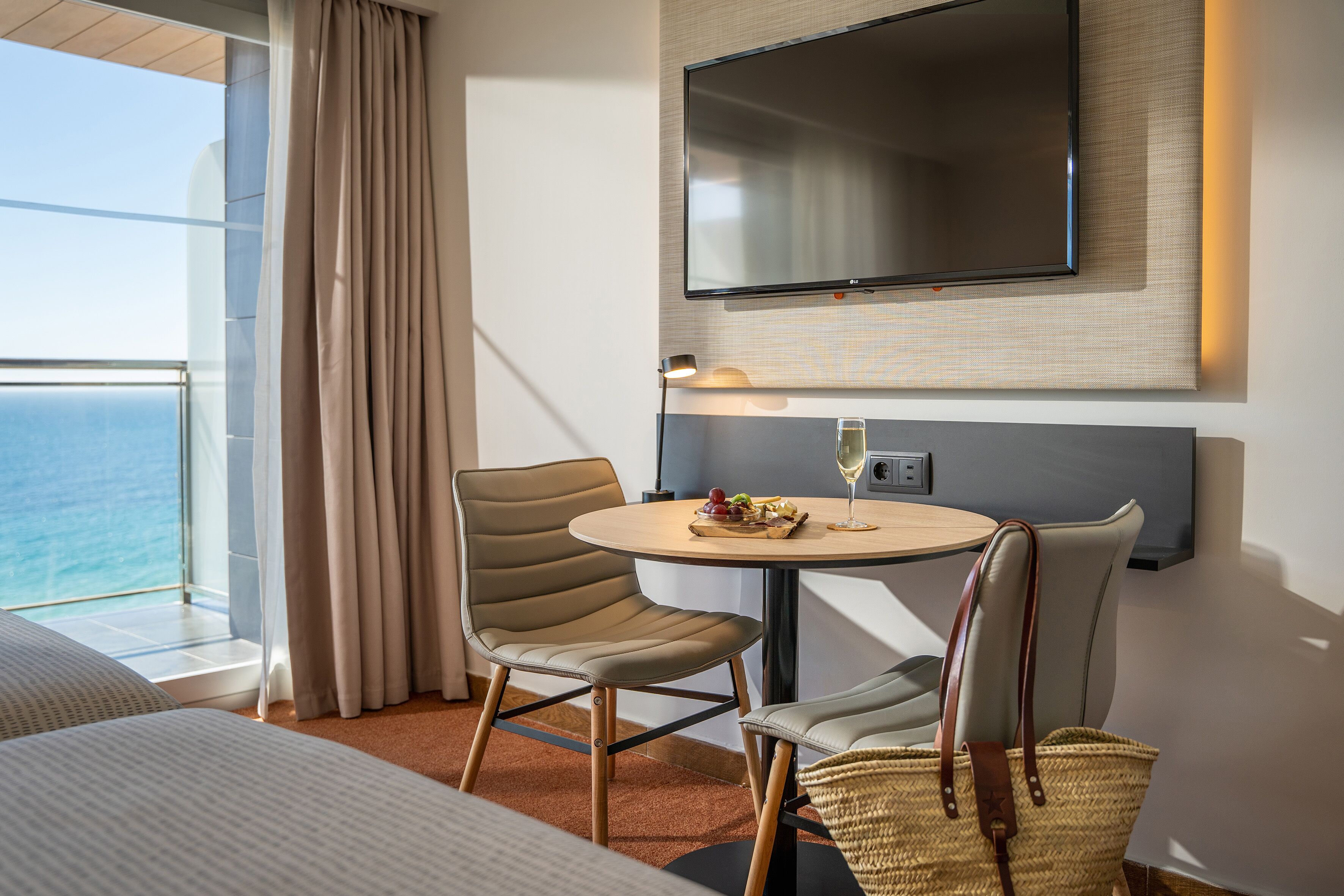 Habitación de hotel con una cama, una mesa redonda con una copa de vino, dos sillas, un televisor montado en la pared y vistas al mar a través de una ventana con cortinas.