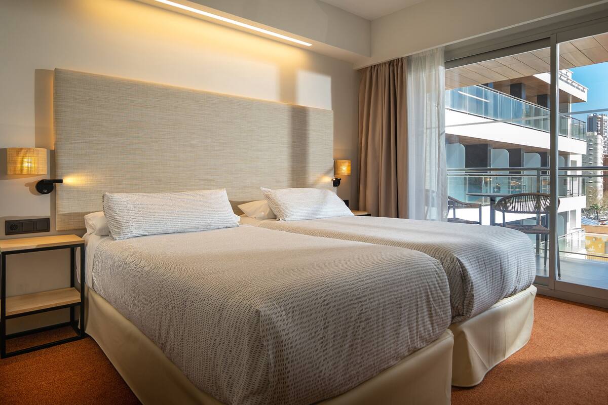 A hotel room with two beds, a large window with a view of the city, and a balcony with outdoor seating.