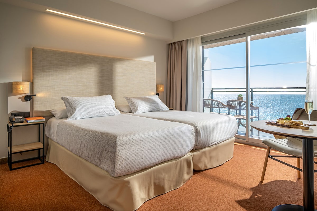 A hotel room with two beds, a small table, a chair, a nightstand, and a large window with a view of the sea.