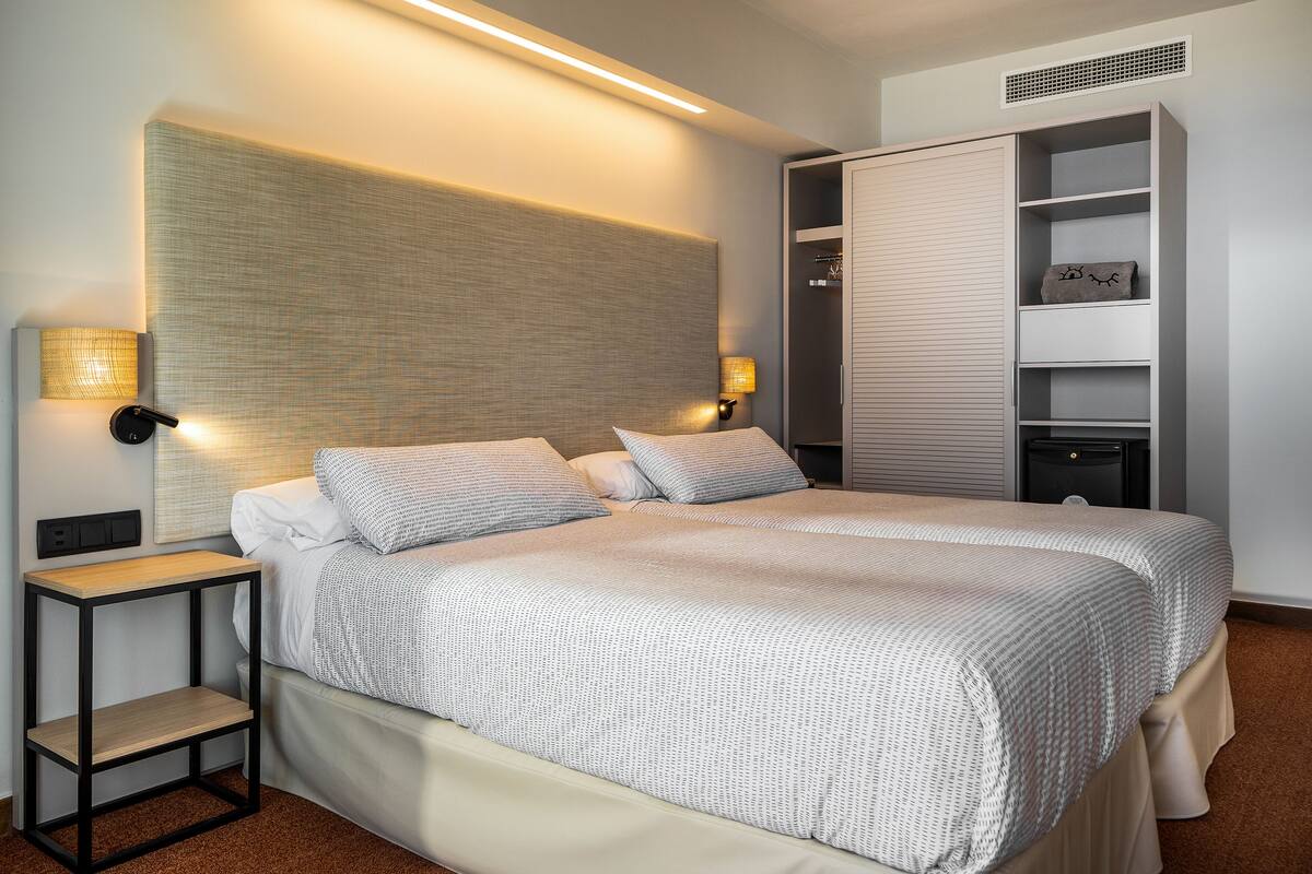 A modern hotel room with a large bed, bedside lamps, a wardrobe, and a small table.