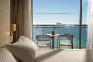 Habitación de hotel con un ventanal grande que da a una masa de agua y una tierra lejana. En el interior, hay una cama con cabecera, una pequeña mesa redonda con dos sillas de mimbre y una lámpara fijada a la pared.