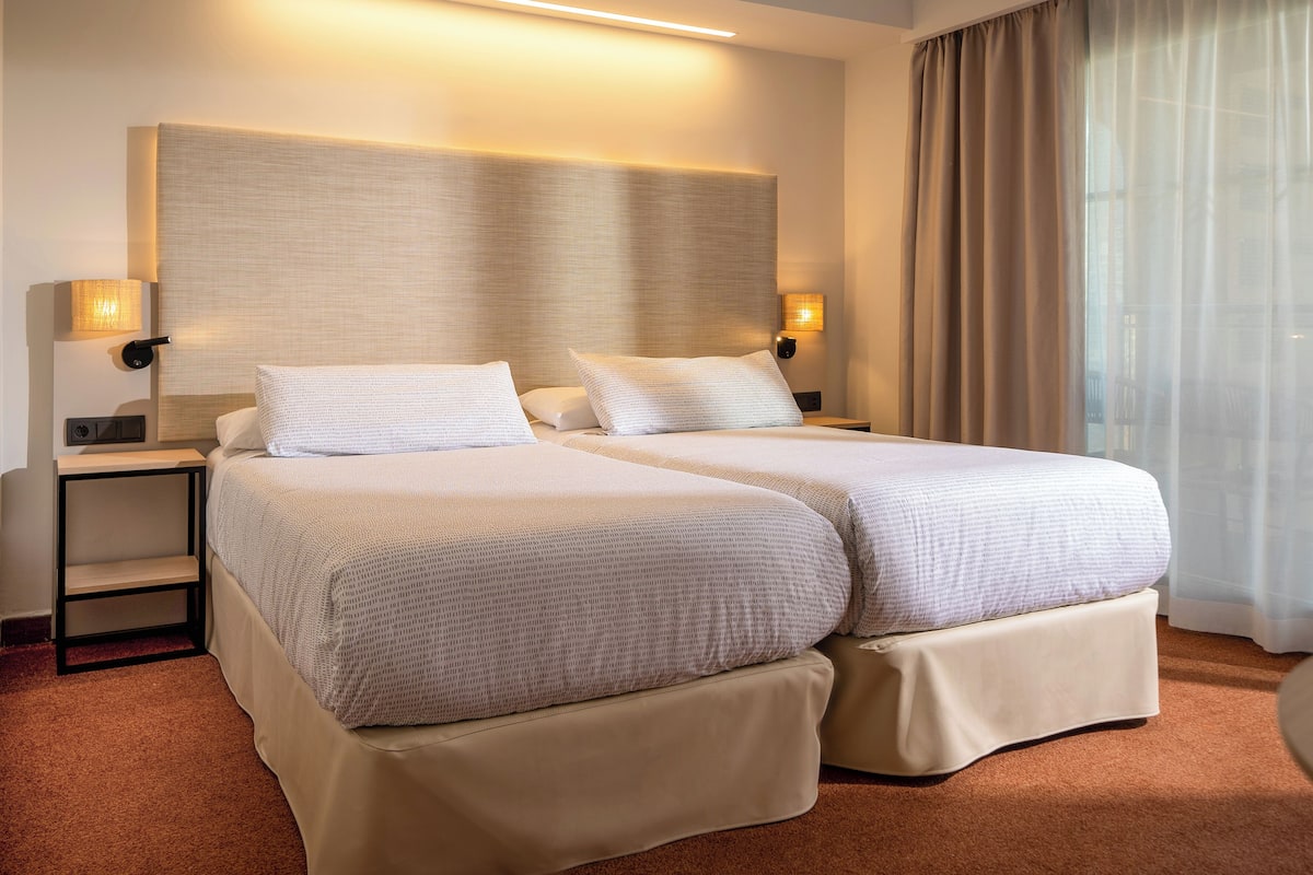 A hotel room with two beds, a large headboard, bedside lamps, and a view of a building through sheer curtains.