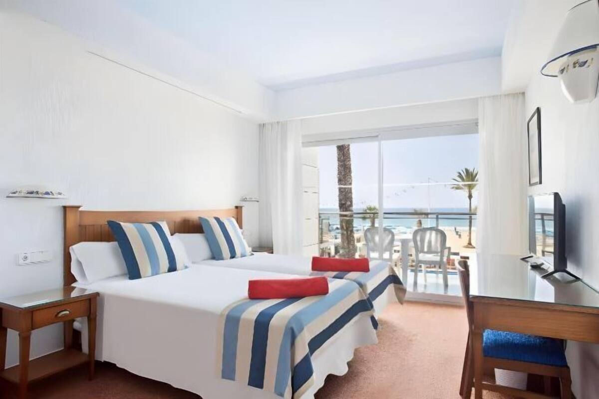 A hotel room with two beds, a desk, and a balcony with a view of the sea.
