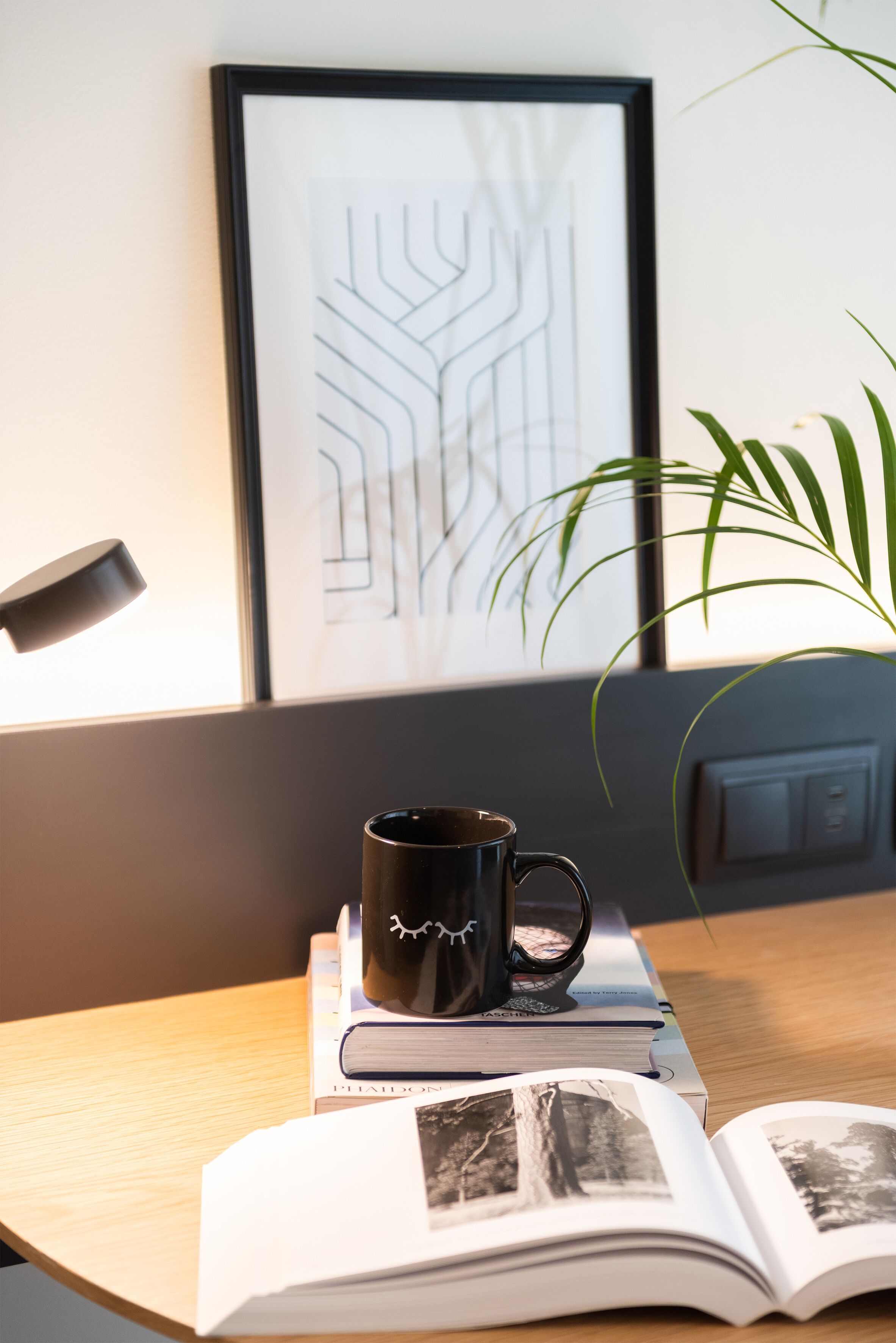A framed abstract artwork, a black mug, and an open book with photographs on a wooden table.