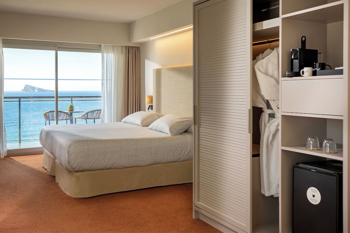 A hotel room with a bed, a small table, chairs, a view of the sea, and a closet with a coffee maker and glasses.