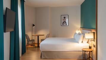 A hotel room with a bed, a desk, a chair, a TV, and a wall-mounted lamp.