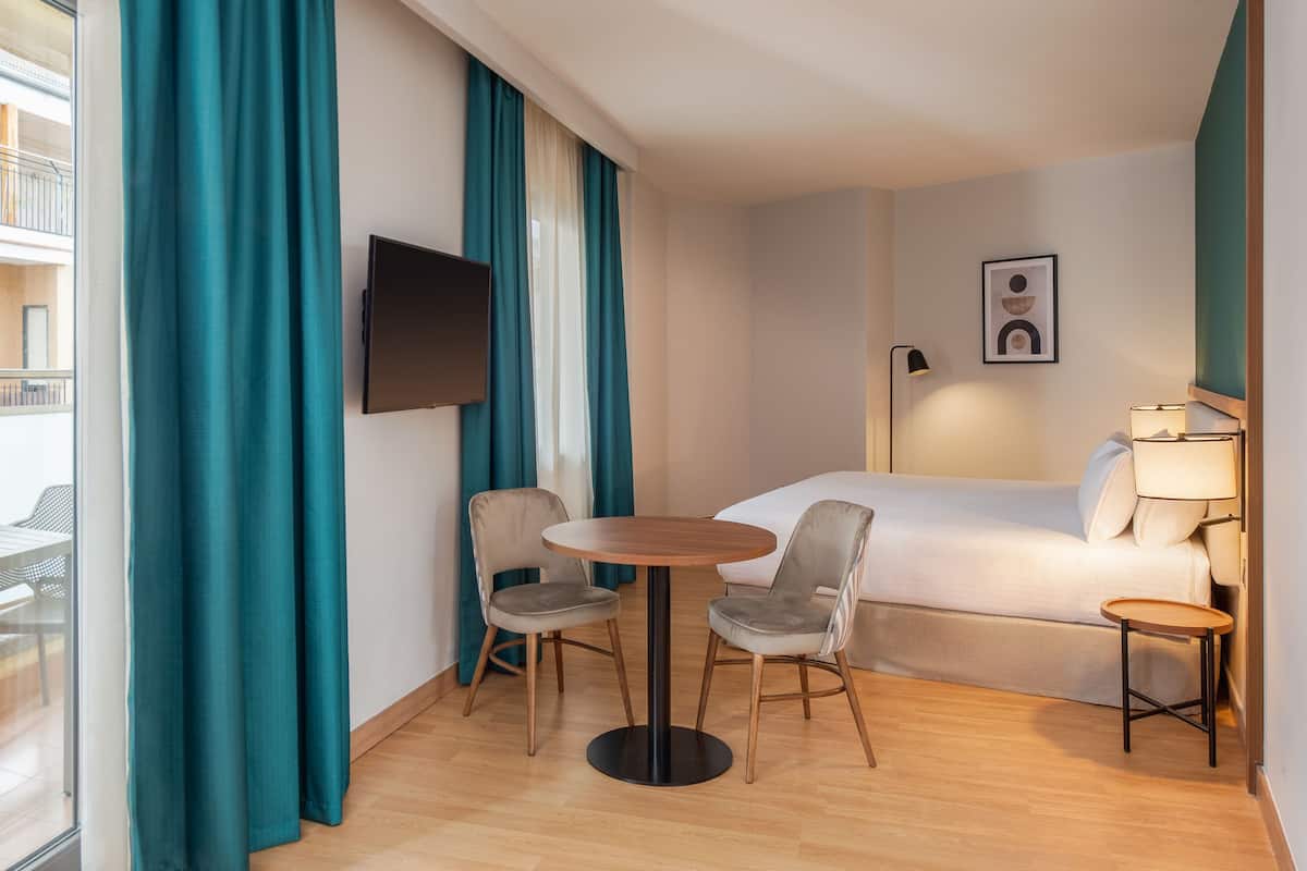 A hotel room with a bed, two chairs, a small round table, a TV, and a balcony with a table and chairs.