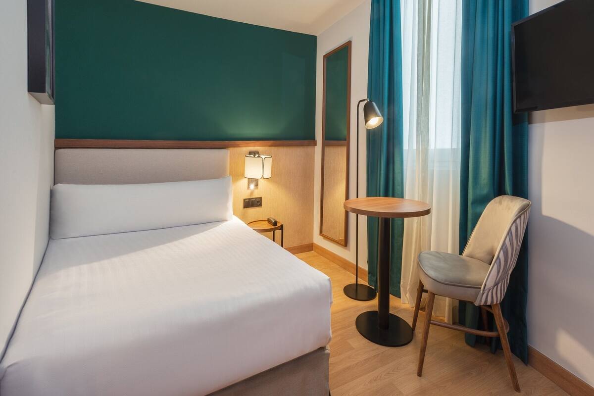 A hotel room with a bed, a chair, a small table, a lamp, and a TV mounted on the wall.