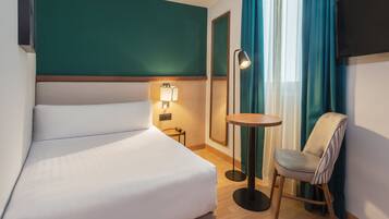 A hotel room with a bed, a chair, a small table, a lamp, and a TV mounted on the wall.