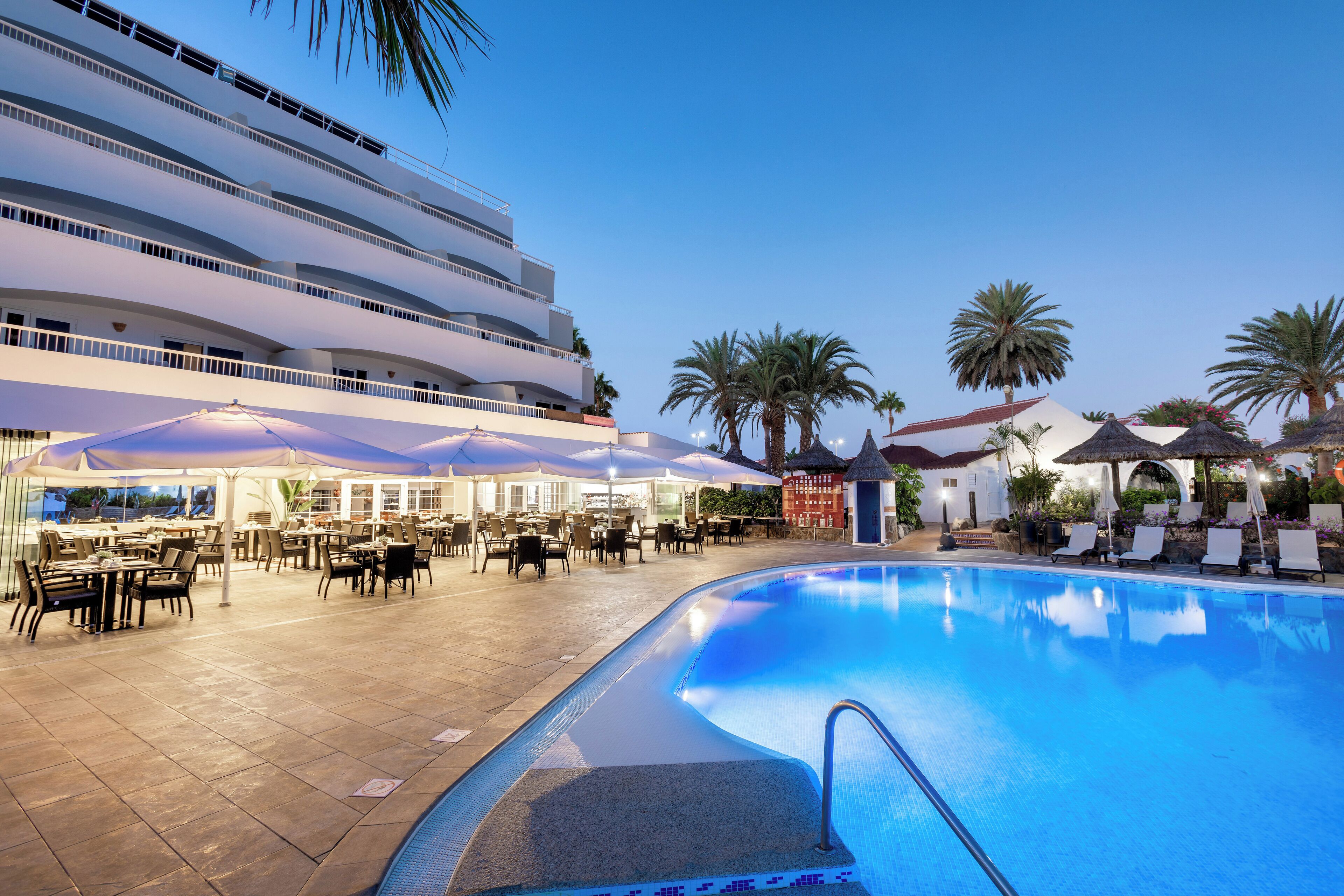 A swimming pool area with lounge chairs and umbrellas, a multi-story building, and palm trees.