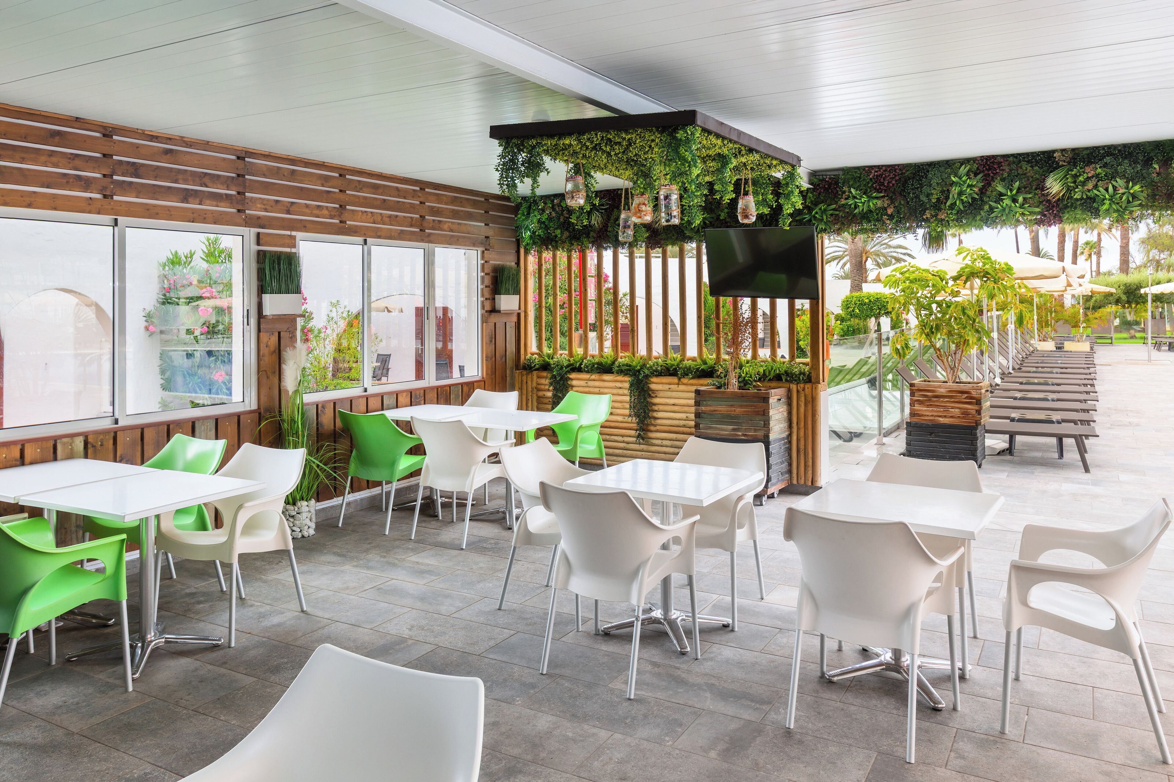 Un café al aire libre con sillas blancas y verdes, mesas de madera y una pared verde con plantas y un televisor.