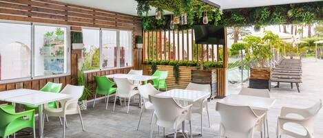 Un café al aire libre con sillas blancas y verdes, mesas de madera y una pared verde con plantas y un televisor.